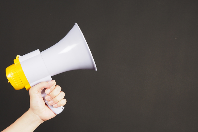 A hand holding a grey megaphone with a yellow trigger against a dark background.
